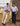 Two men standing outdoors in a grassy field next to a tree, wearing light blue long-sleeve button-up shirts and different pairs of jeans, one in beige and one in dark blue. A brown fabric backdrop is visible on the right.